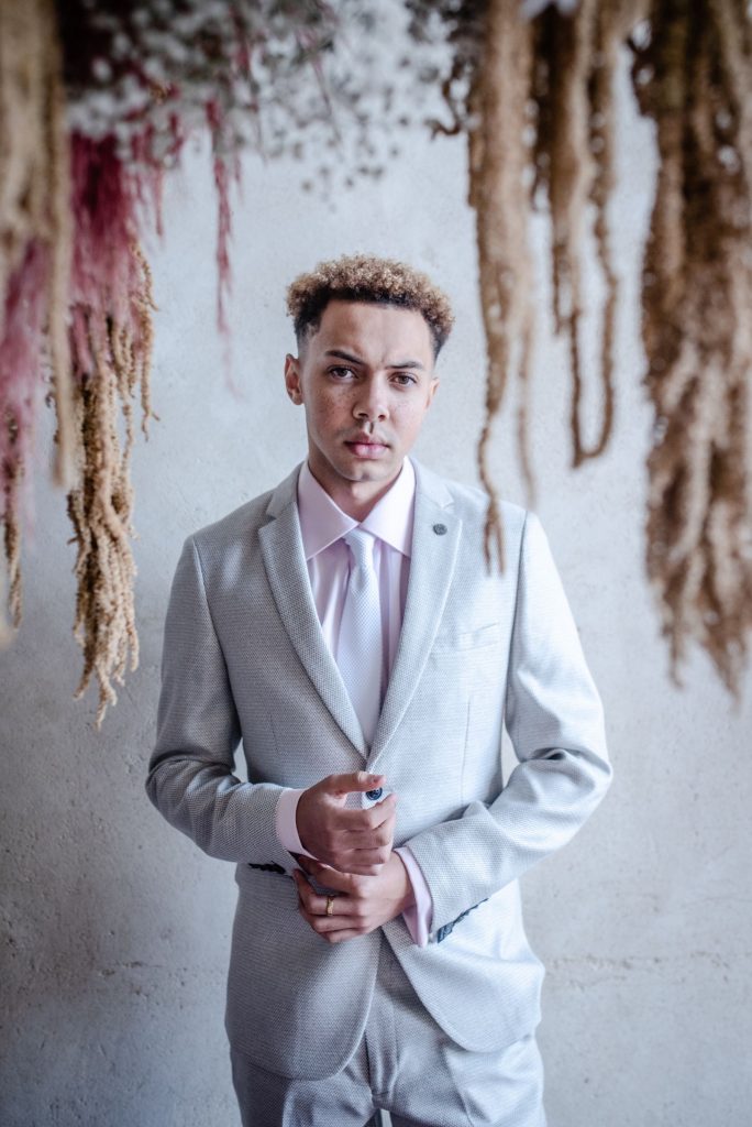 A cool, modern groom wears a light grey suit and tie with a light pink shirt. He stares into the camera.