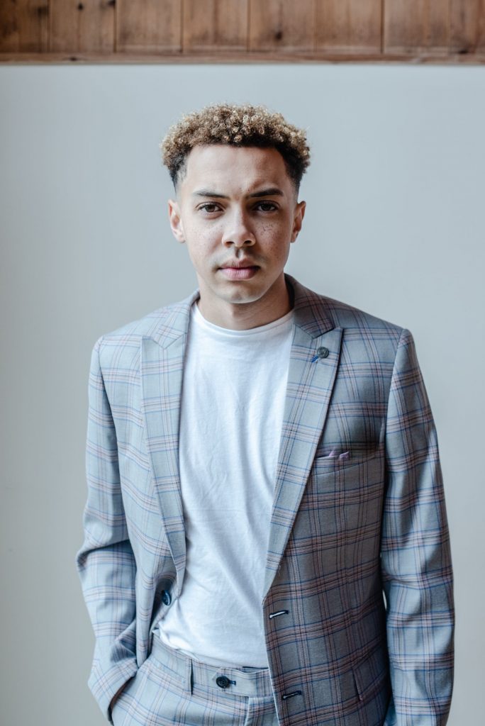 A young groom is dressed in a contemporary style wedding outfit with a grey checked open buttoned suit jacket underneath which he wears a white t-shirt.