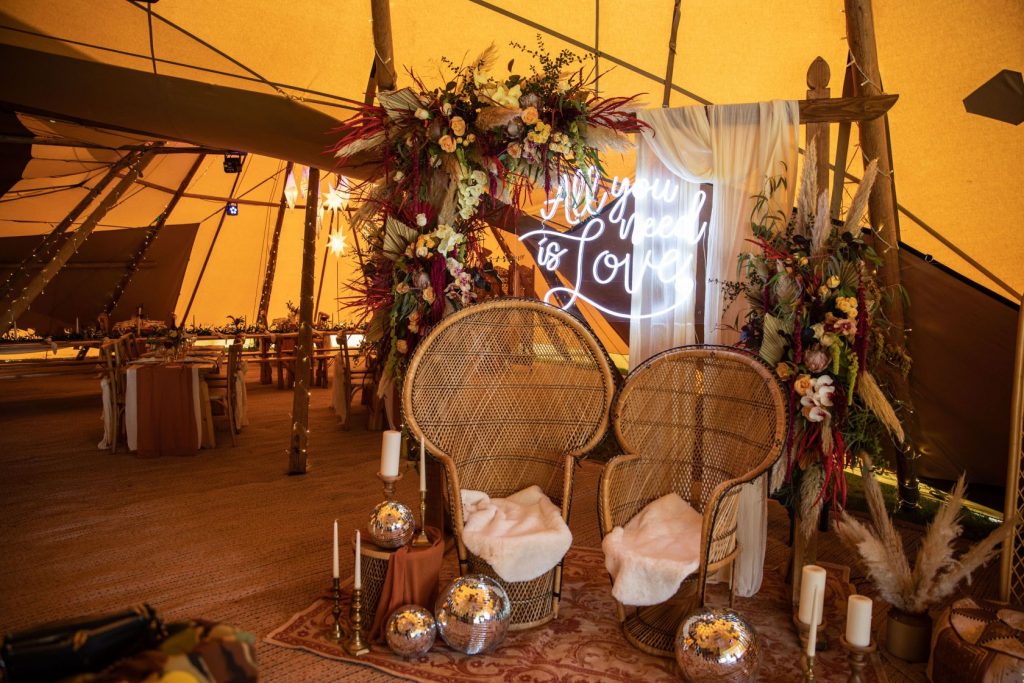 Two rattan peacock chairs are styled for a boho style wedding shoot complete with a flower arch backdrop with a hanging neon sign and a selection of glitter disco balls and candles on the floor