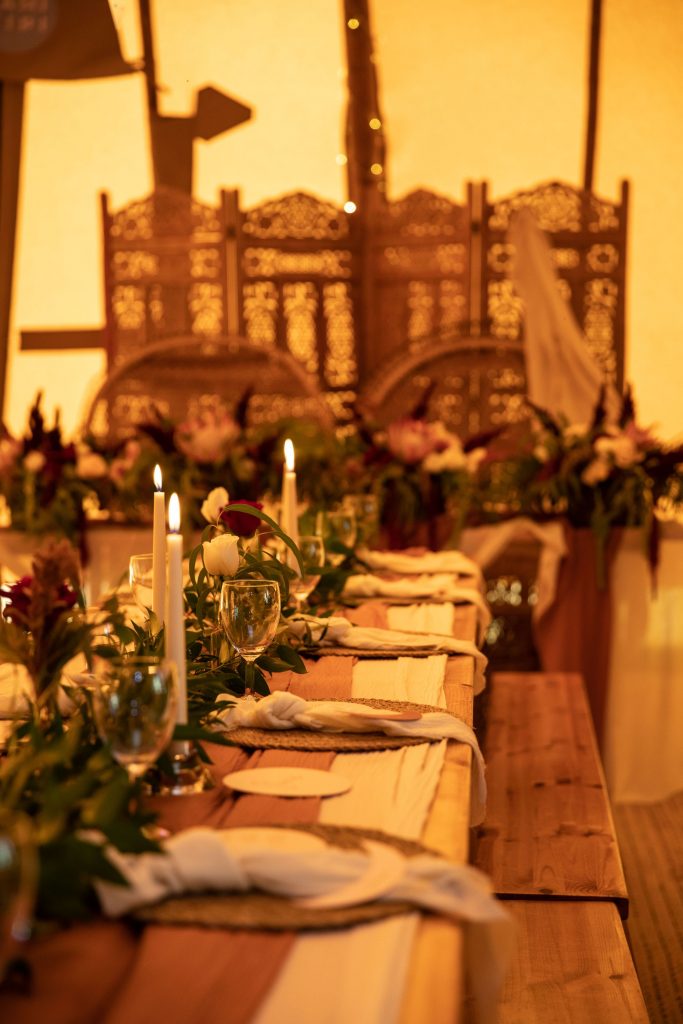 A rustic style long wooden wedding table is set up inside a tipi tent for a boho style wedding. The table is decorated with simple candles and small red flowers with masses of green foliage.