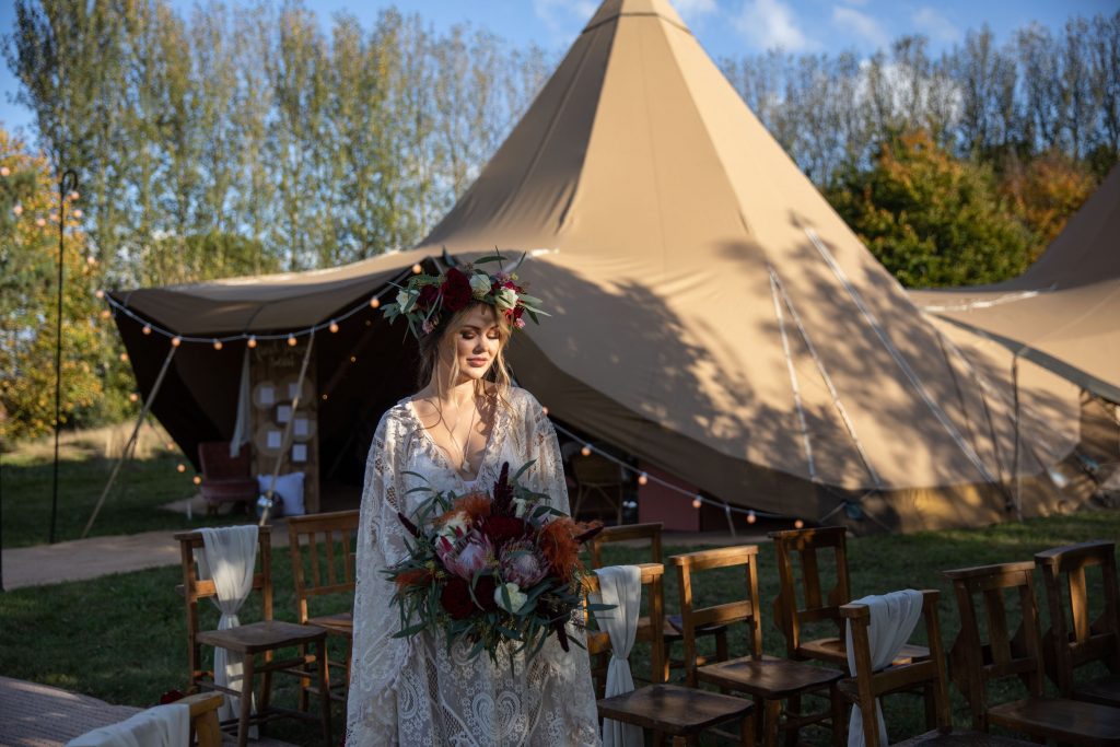 A boho style bride stands holding a large wedding bouquet of red and white flowers with green foliage. She wears a fresh flower floral crown to match. Behind her is a large tipi in which her wedding is taking place.