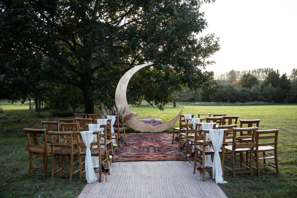 An outdoor ceremony set up with wooden chairs in rows. The end chair on each row has a white sash tied to it and the aisle floor is lined with a vintage red rug. At the front of the ceremony space is a large crescent moon backdrop.