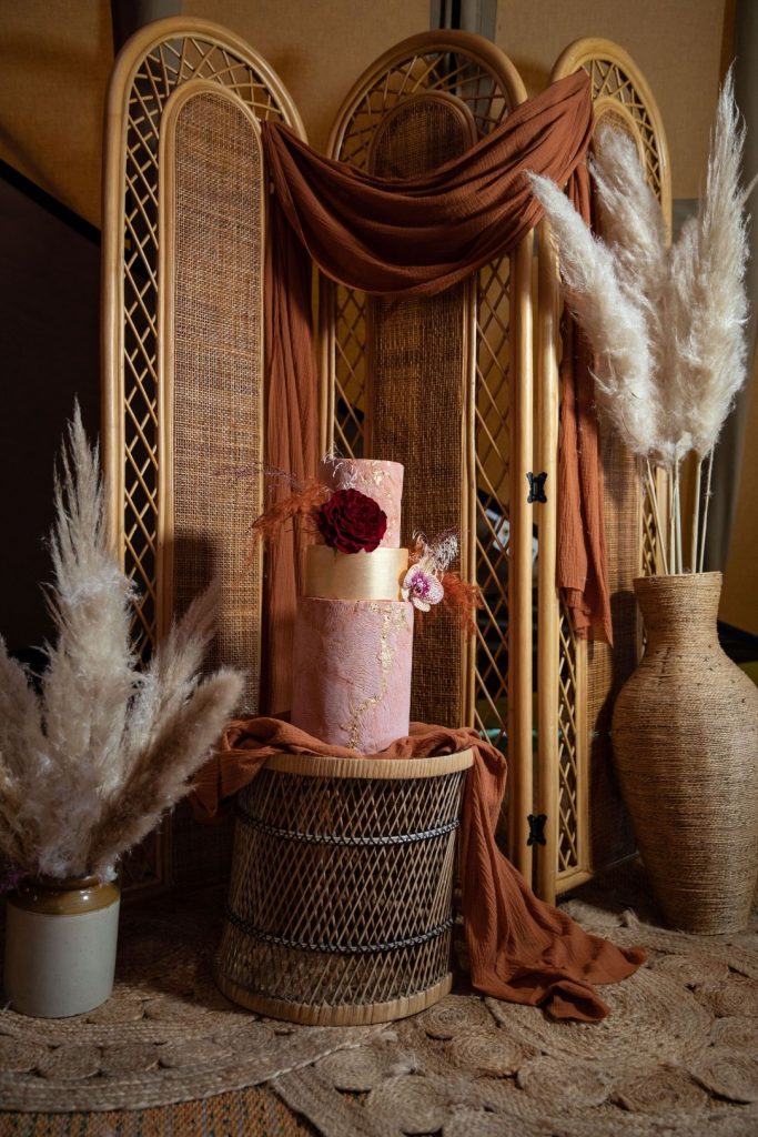 A two tiered pink terracotta wedding cake with hints of gold and bright deep red flowers sits as a cake display in front of oversized vases filled with pampas.