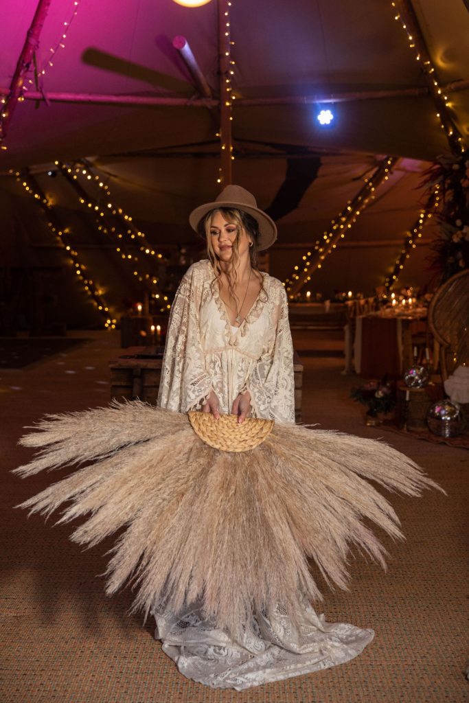 A boho bride wears a lace wedding dress and a brown Fedora hat. She holds a large pampas fan in her hands.