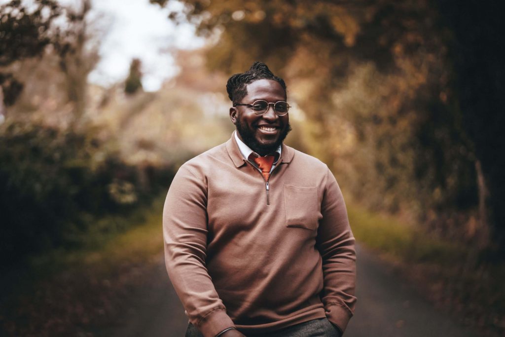 A smiling man looks towards the camera as he walks though a clearing in a forest. He wears glasses and looks smart in a zip down light brown jumper and tie. He has one hand in his trouser pocket.