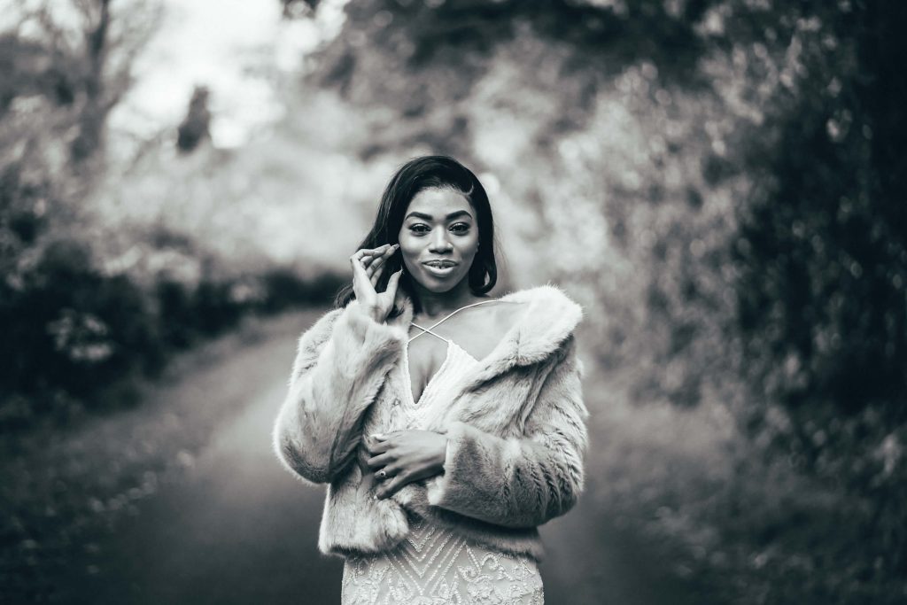 Beautiful black and white image of a woman wearing a glamourous sparkly white dress with a faux fur jacket on top. She holds one arm across her body whilst the other gently moves her hair from her face.