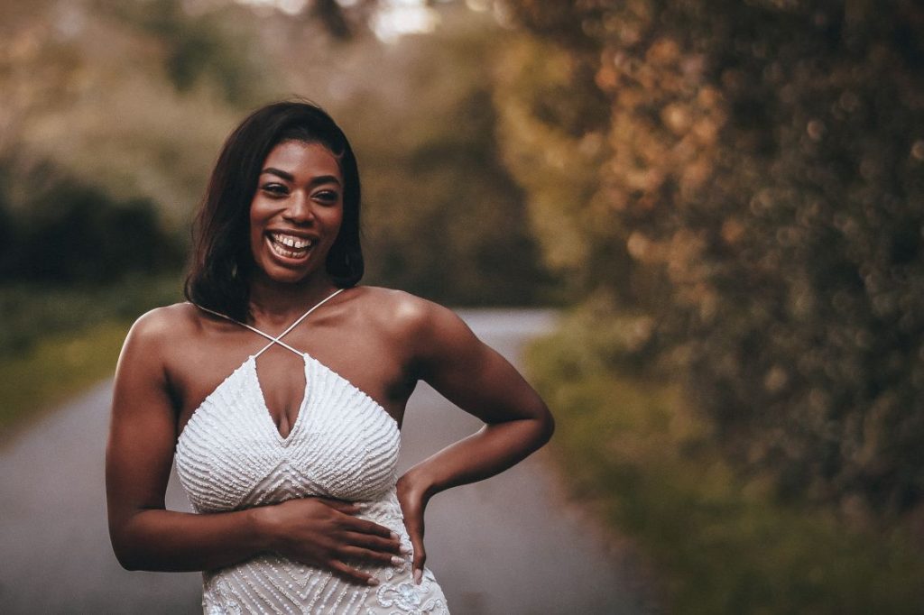 A bride smiles broadly as she poses during her intimate wedding photo shoot. She wears a glamourous white sparkly dress with a keyhole halter neck. One hand is on her hip whilst the other she holds across her stomach.