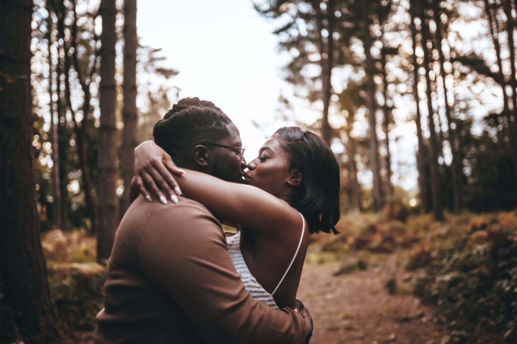A couple embrace in the woodlands. They gently kiss as the woman wraps her arms around her husband's neck.