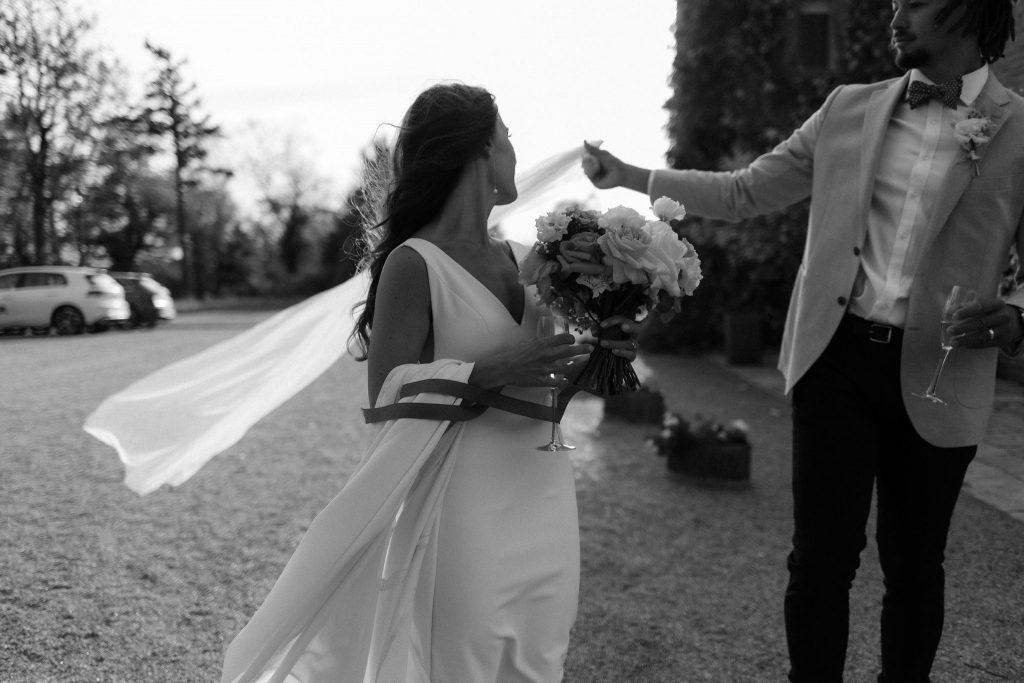 A bride's veil comes loose from her hair, and her groom grabs it and holds it for her as it blows in the wind. The bride looks elegant in a minimalistic sleevless wedding dress as she looks behind her to see what her groom is doing. She holds a beautifully elegant bouquet of roses.