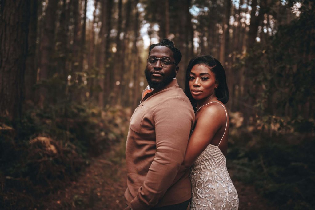 A bride and groom stand together, the groom has his back to his new wife as she puts her arms around his waist. They both look straight into the camera. The setting is a clearing in a forest.