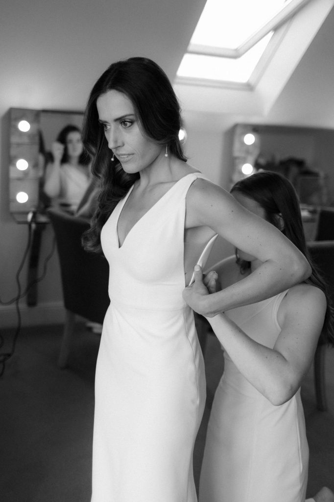 A bride is helped into her wedding dress by a bridesmaid. The wedding dress is a sleeveless simple unfussy dress which is refined and elegant.