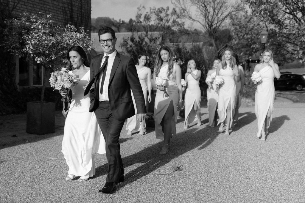 A black and white image of a bride walking arm in arm with her father on her way to her wedding ceremony. Her bridesmaids who wear matching dresses walk behind them.