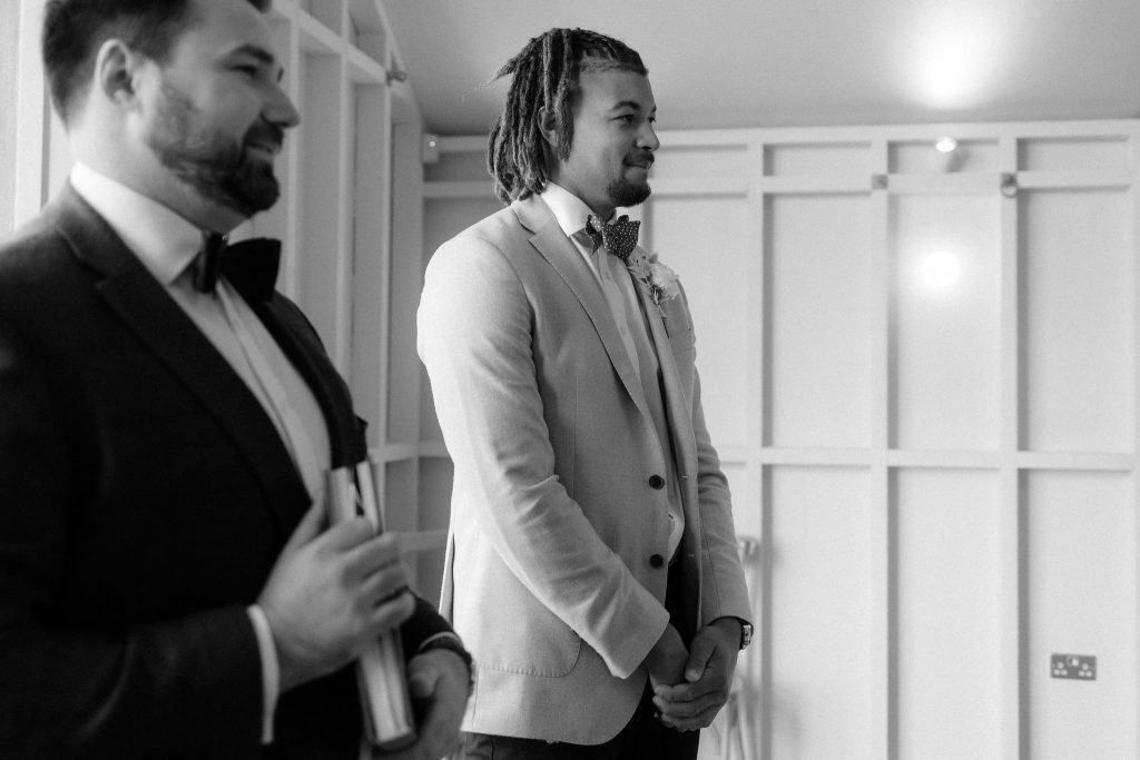 An emotional looking groom waits at the front of his ceremony room for his bride to arrive. He clasps his hands together, wearing an open jacket with a bow tie.
