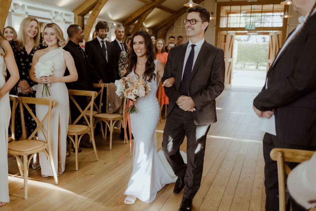 A bride walks down the aisle in her contemporary barn wedding venue at Hyde House, arm in arm with her father. They both smile broadly.