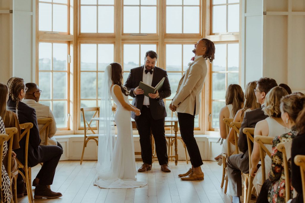 A bride and groom captured mid way through their wedding ceremony which is being conducted by the Grooms best man. The groom throws his head back in laughter as something funny is being said.