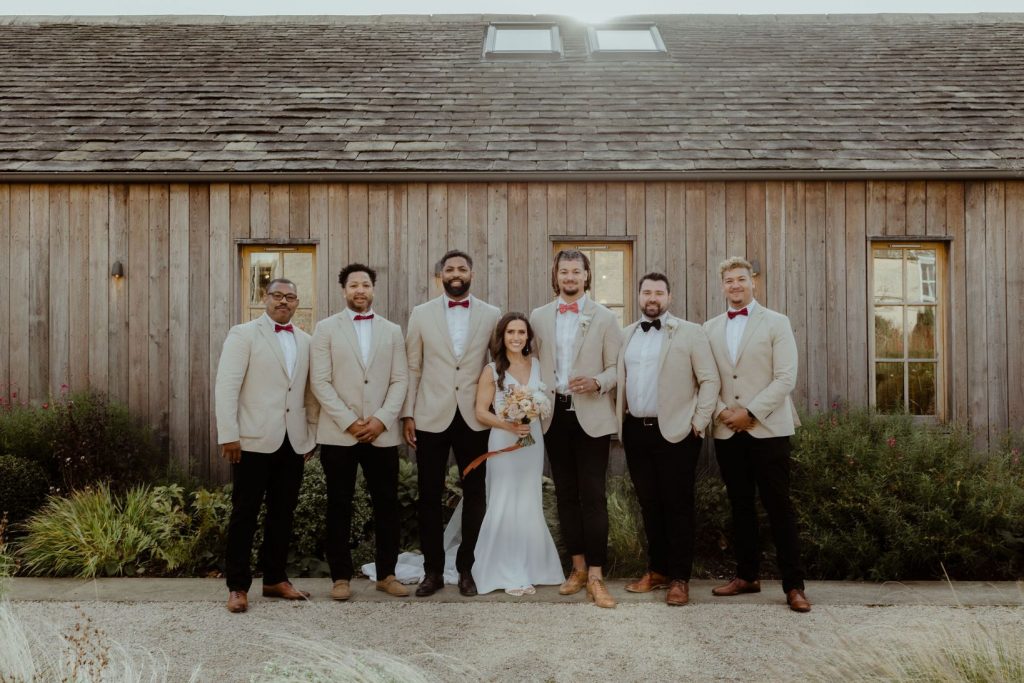 A group wedding shot of a bride and groom standing in the centre of the 5 groomsmen. The men all wear smart casual beige jackets and bow ties. The bride wears a simplistic yet elegant wedding dress and holds a beautiful bouquet of toffee and cream flowers. at the beautiful wedding venue of Hyde House.