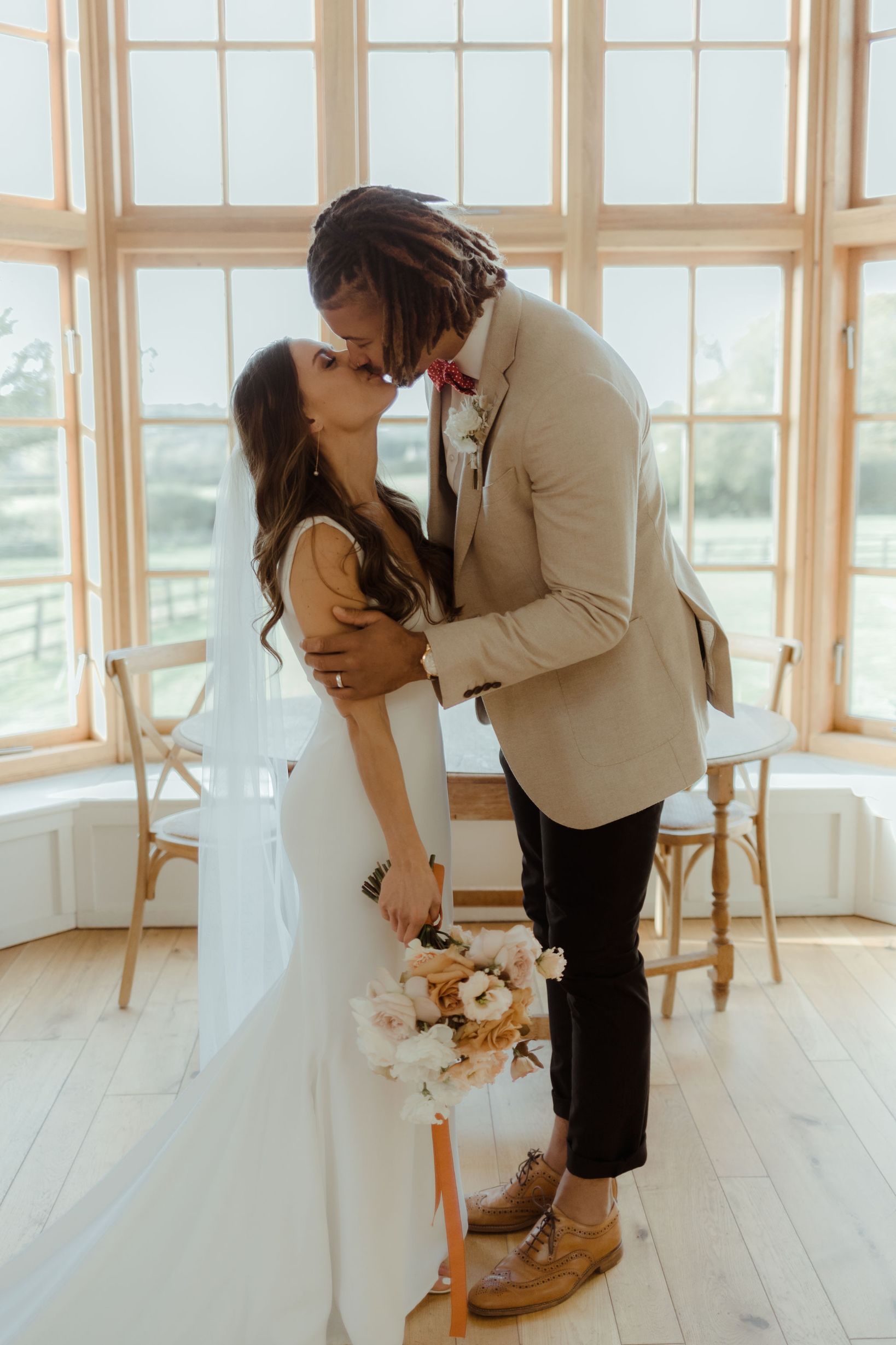 A Groom kisses his new wife on the lips and gently holds her arms as they embrace. The bride holds a toffee and cream coloured bouquet.