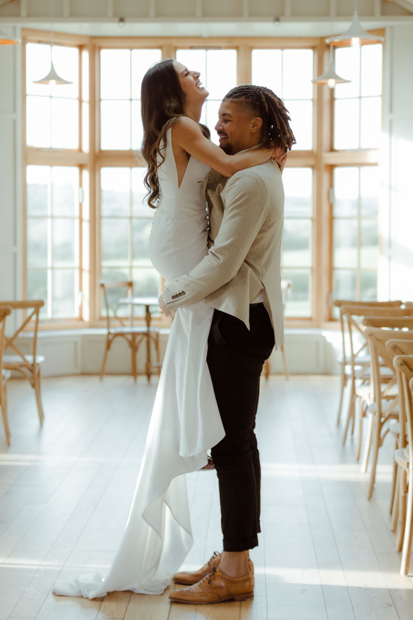 A groom is lifting up and hugging his new bride. They embrace in a light and airy ceremony room with a large window behind them which lets in lots of natural daylight.