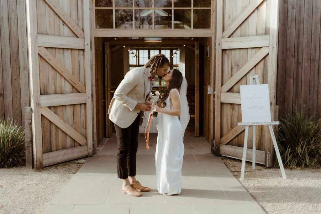 A tall groom bends down to kiss his new wife as they stand in front of their ceremony room at Hyde House in the Cotswolds.