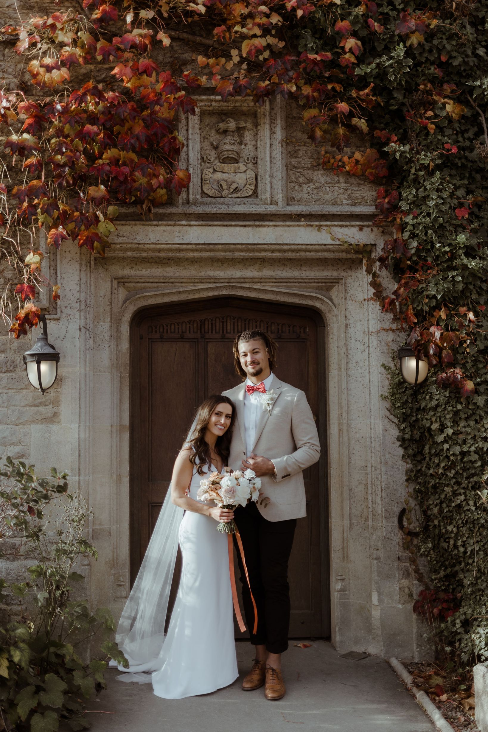 A bride and groom stand in front of a doorway outside a building. The exterior of the stone walls is covered with red and green leaves.