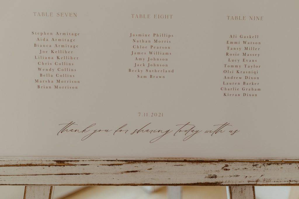 A minimalistic wedding table plan of dark plain writing and calligraphy font on a white card which sits upon a wooden easel at Hyde House in the Cotswolds.