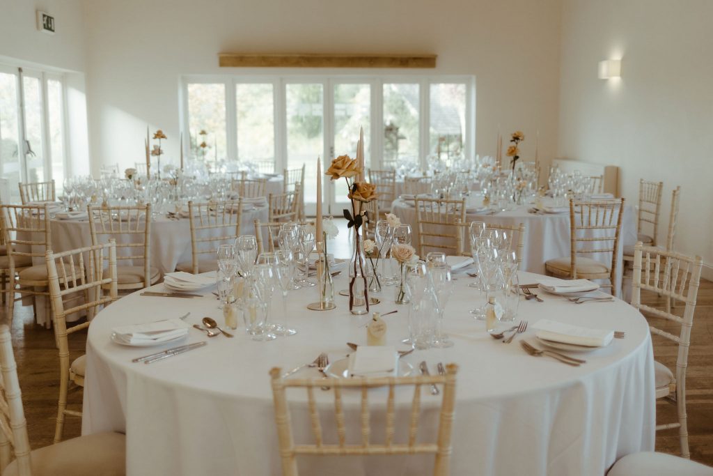 An elegant wedding display of white clothed round tables and wooden chairs. The tables are decorated with simple clear jars with toffee coloured roses and white ranculus. Lots of natural light streams through into the wedding room at Hyde House giving a light and airy feel.