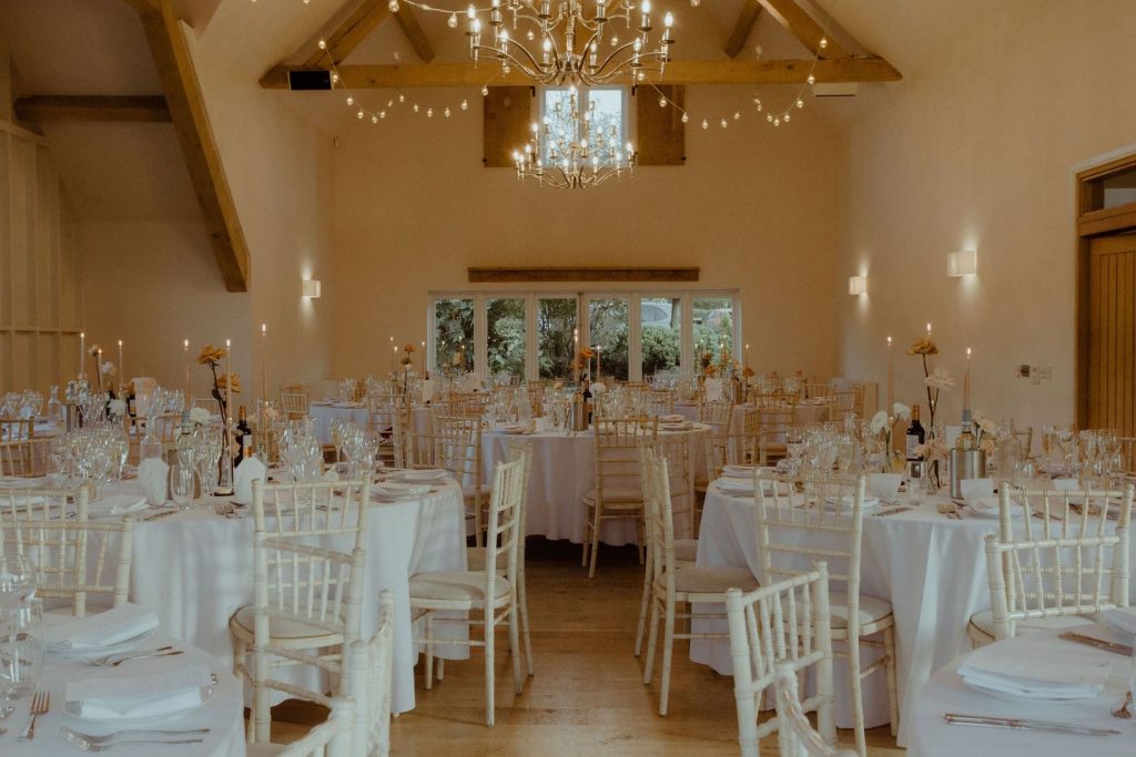An elegant, refined wedding set up inside Hyde House wedding venue. The round clothed tables are decorated simply with jars of single stems of toffee colour roses and white ranculus flowers. From the ceiling, strings of fairy lights hang overhead.