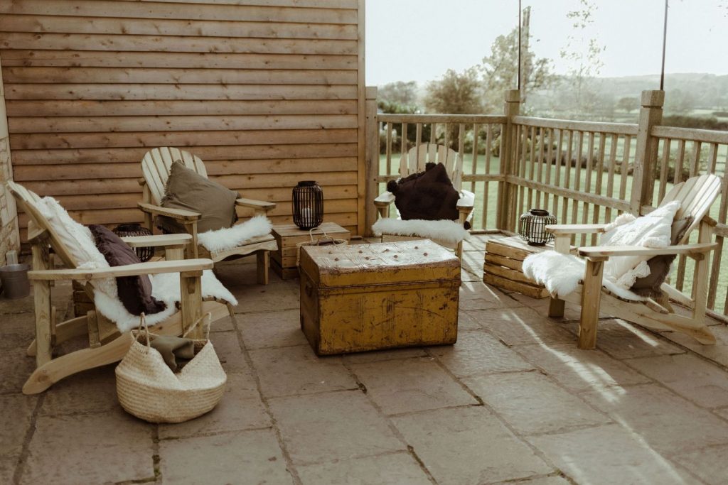 An outdoor chill out area with wooden furniture and cushions is set up ready for a wedding party at Hyde House in the Cotswolds.