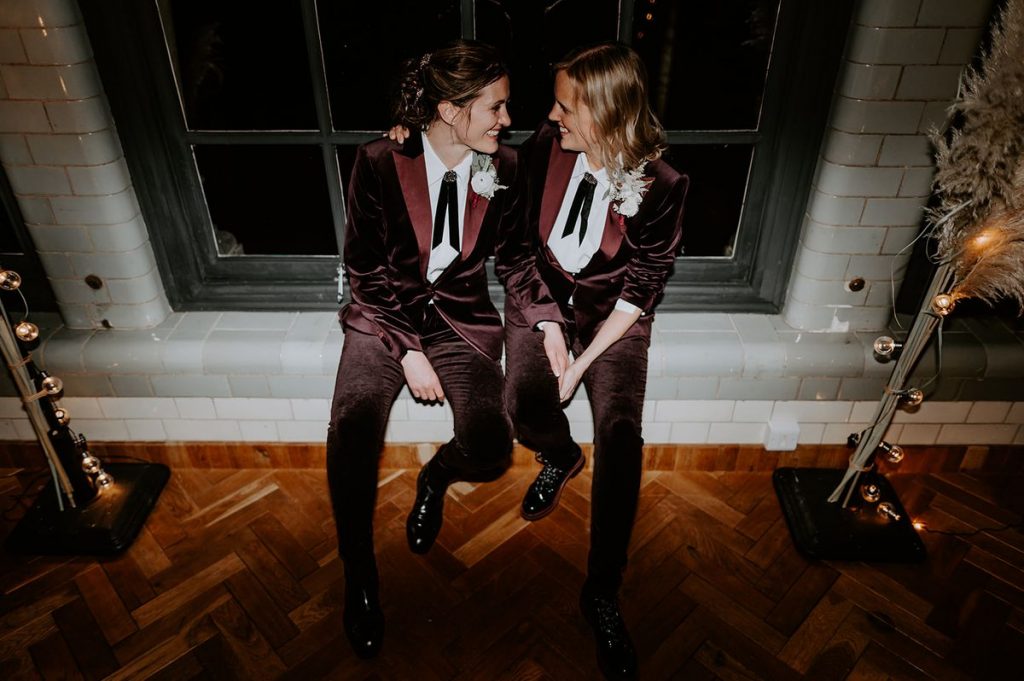 Two brides sit on a windowsill at The Pumping House during the evening part of their wedding day. Previously they were both wearing wedding dresses but have changed into matching plum velvet suits with small black ties and a small white flower. They look uber-cool as they casually chat to one another.
