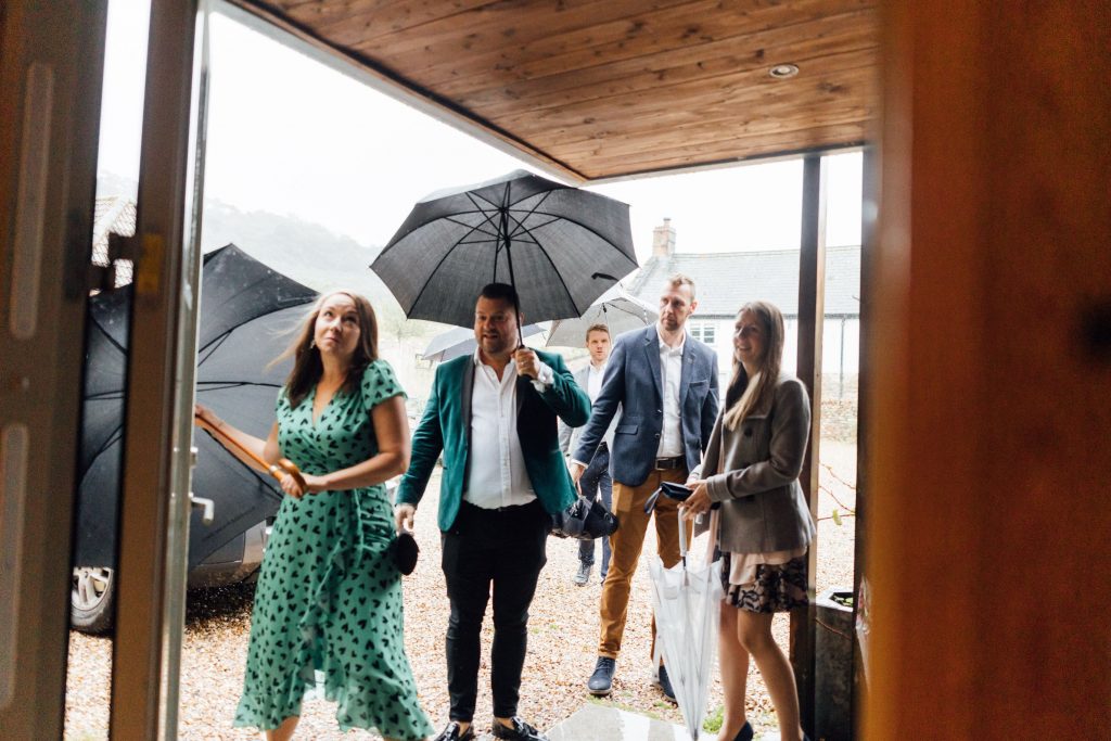 Wedding guests arrive at River Cottage for a non-traditional wedding day holding umbrellas over head to shield from the typical rainy English weather. Despite the rain, all the guests are smiling and happy.