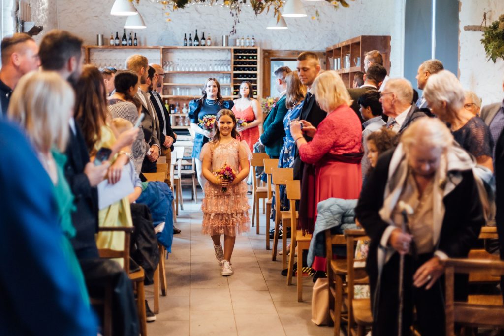 A young bridesmaid in a non-traditional bridesmaid outfit walks up the wedding aisle at River Cottage