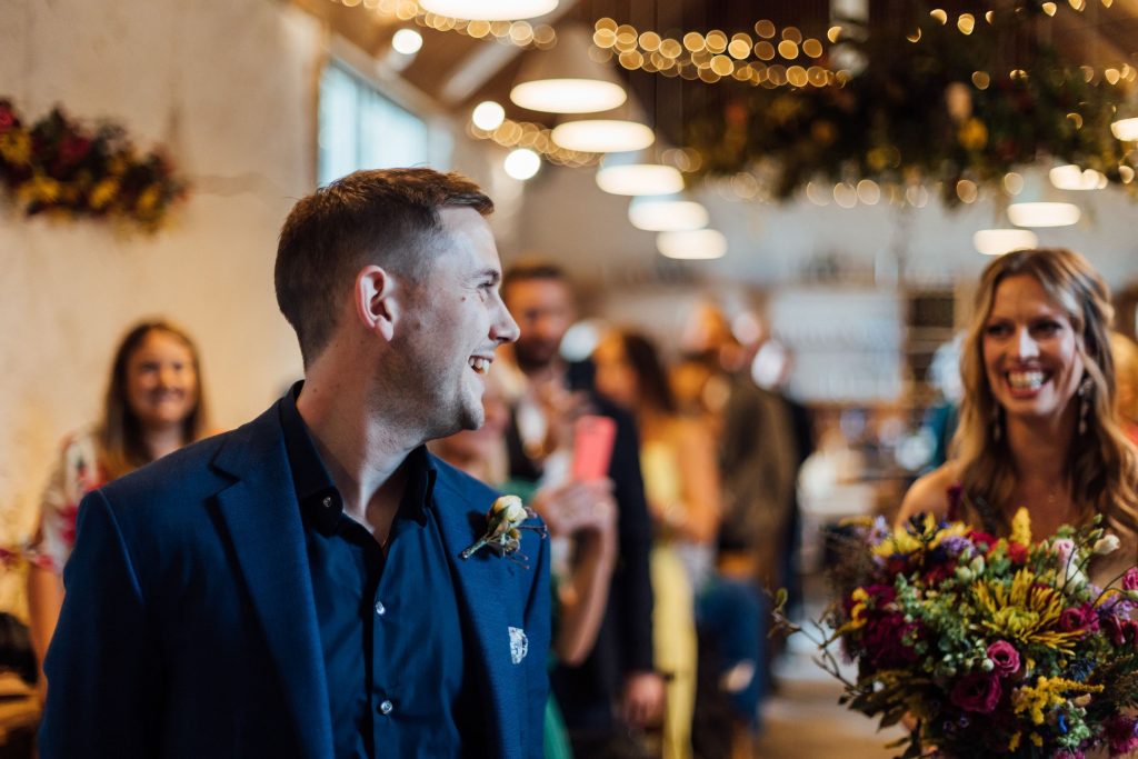 A groom wearing a casual blue suit and open collar blue shirt turns to look down the aisle at his soon to be wife. His mouth is open wide and he is smiling broadly. The bride is looking towards her soon to be husband with the biggest of grins, showing pure excitement.