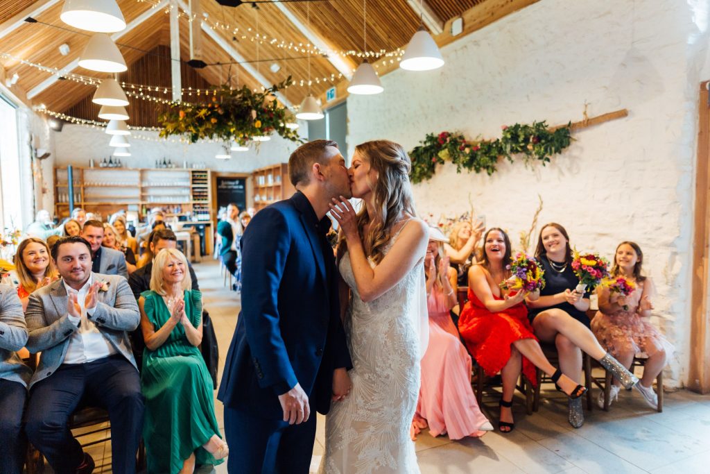 A newlywed couple kiss during their wedding ceremony. The stand in front of their guests who are seated and all smiling with pride. The bride cradles her new husbands face in her hands.