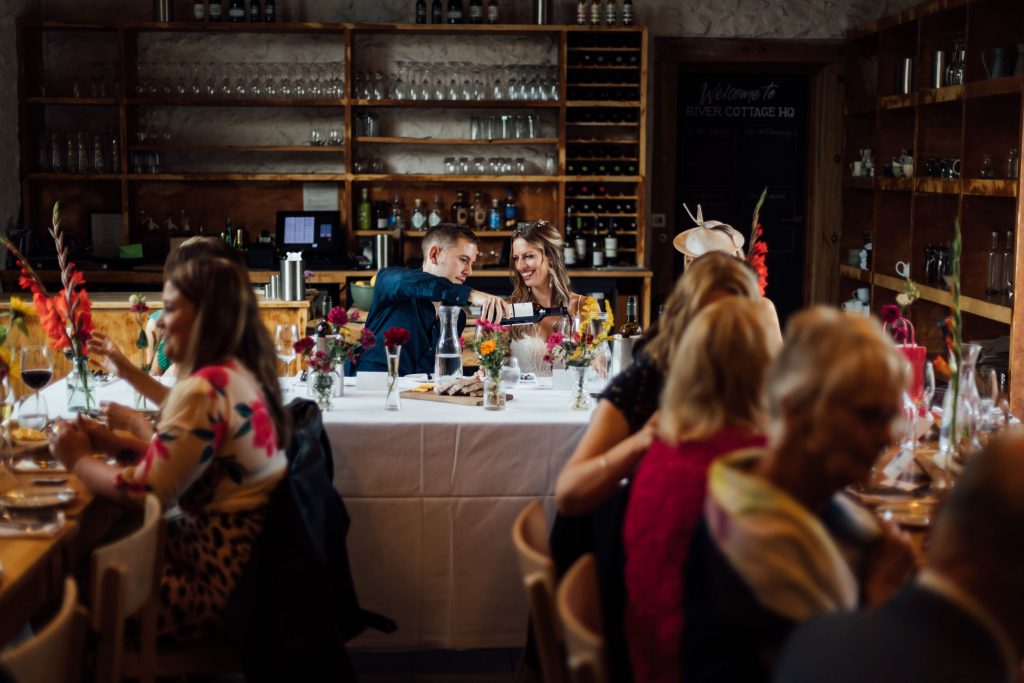 A beautiful capture of a bride and groom seated at their wedding table during their non-traditional wedding celebrations. The groom is pouring a glass of wine for his new wife as she smiles broadly. Guests ca be seen in the forefront, but the focus of the image is directly on the newlyweds who seem to be in their own bubble of bliss, oblivious to what's happening around them.