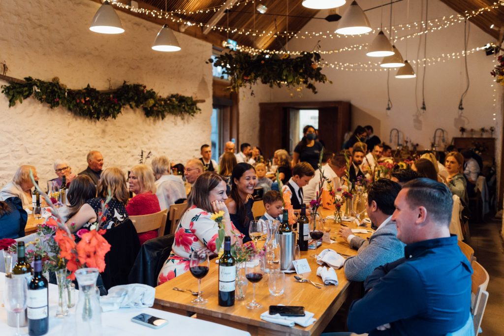 Jubilant candid scenes from a wedding breakfast speech with guest sat around long wooden tables. The guests are all happy and cheering.