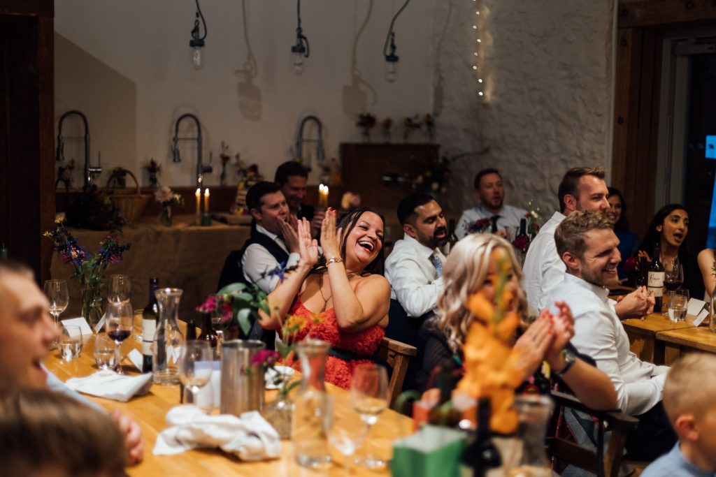 Jubilant candid scenes from a wedding breakfast speech with guest sat around long wooden tables. The guests are all happy and cheering.