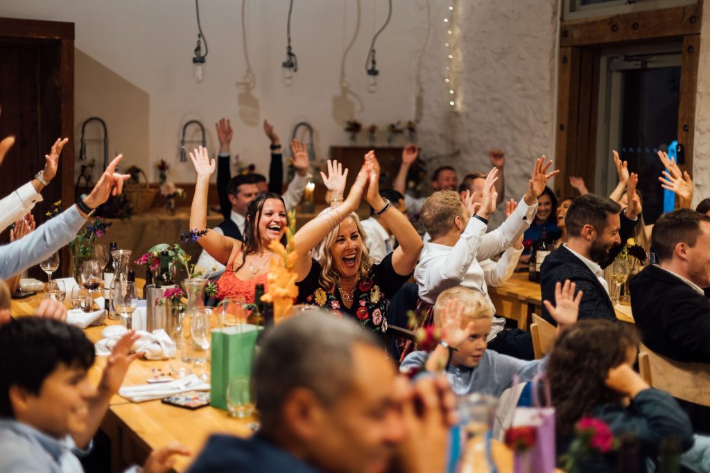 Jubilant candid scenes from a wedding breakfast speech with guest sat around long wooden tables. The guests are all happy and cheering.