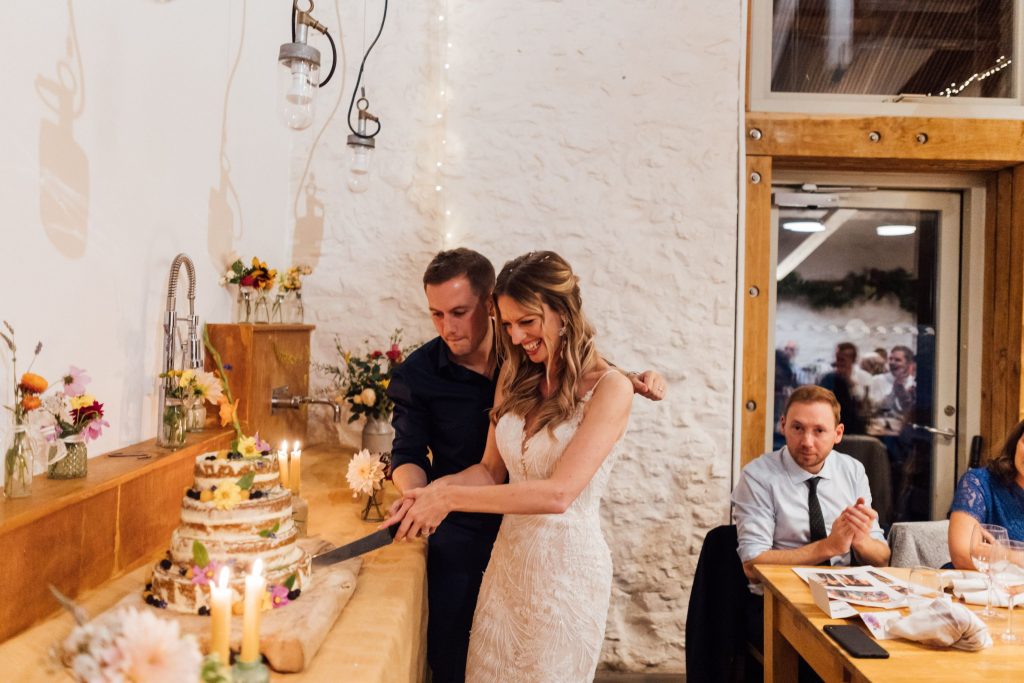 A bride wearing a long white dress stands next to her husband who wears a casual blue suit and dark blue open collared shirt as they hold a knife together as they cut their semi naked wedding cake together.