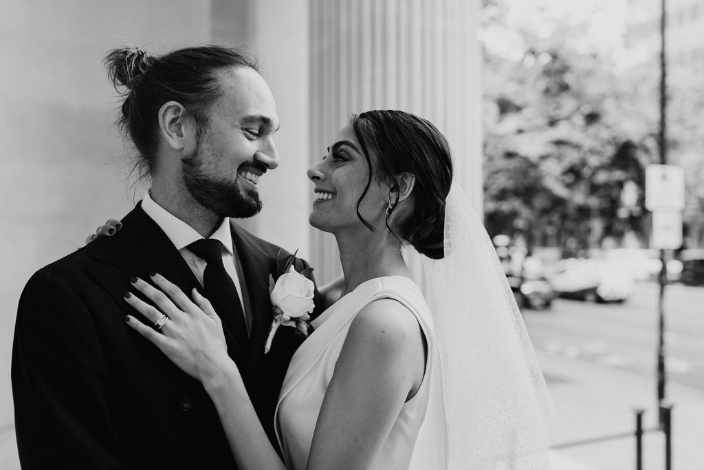 A bride and groom stand facing one another with a complete look of love and adoration for one another as they look into each other's eyes and smile. The bride wears a short polka dot veil in her hair and the groom wears a smart dark jacket and tie with a white shirt. He has a large single white rose button hole.
