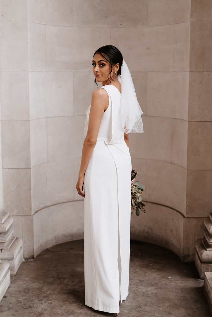 A bride stands outside the Old Marylebone Town Hall posing in her wedding day outfit. She wears a white Halston bridal jumpsuit with one shoulder and holds a small green and white wedding bouquet. In her hair which is swept into a low bun with soft loose curls framing her face, she has a short polka dot white veil.