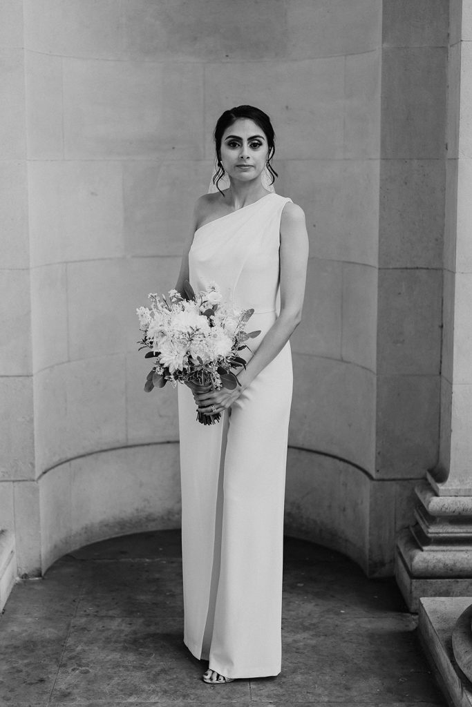 A bride stands outside the Old Marylebone Town Hall posing in her wedding day outfit. She wears a white Halston bridal jumpsuit with one shoulder and holds a small green and white wedding bouquet. In her hair which is swept into a low bun with soft loose curls framing her face, she has a short polka dot white veil.