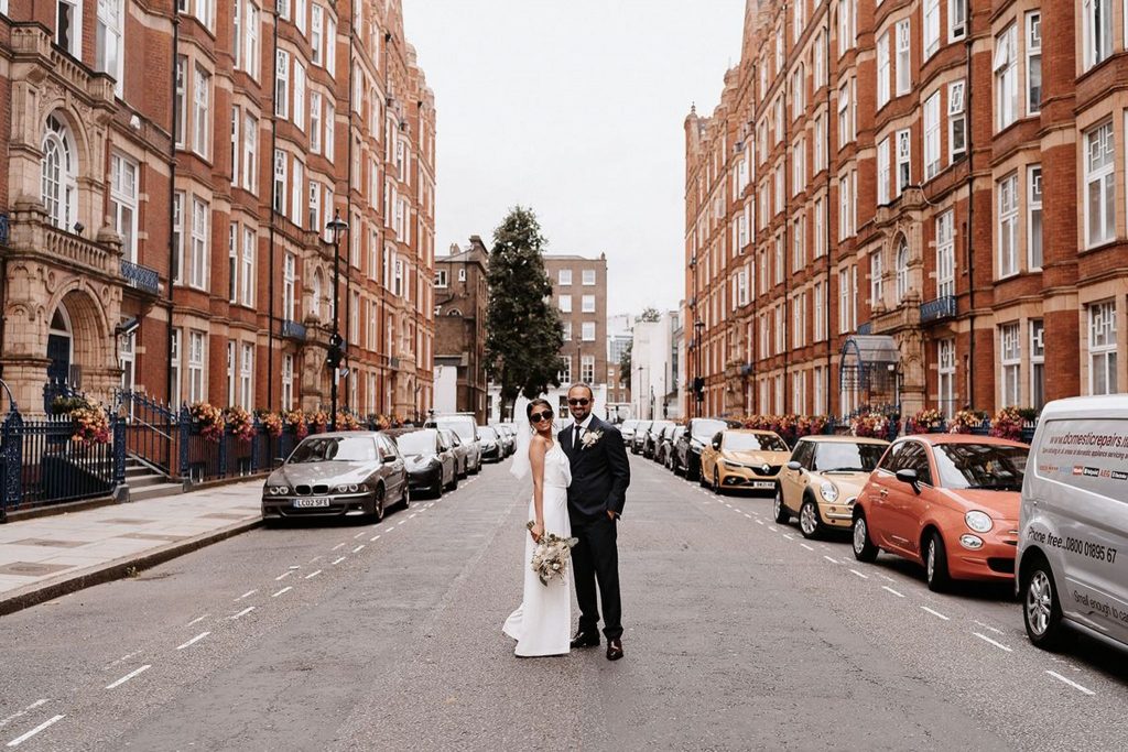 A bride and groom stand posing in the middle of a London road. Cars line the road on either side and tall office style buildings can be seen on either side of the road. The couple look very cool wearing black shades and the bride wears a white bridal jumpsuit.