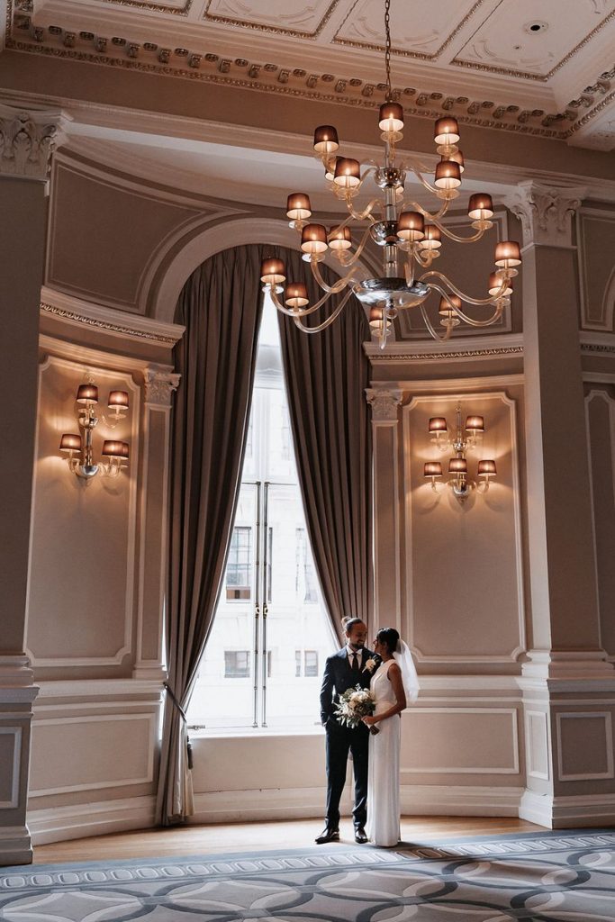A bride and groom stand next to a huge floor to ceiling window with sumptuous curtains and an elegant chandelier. This is one of the grand rooms in the luxurious Corinthia London where the couple are hosting their intimate wedding celebrations.