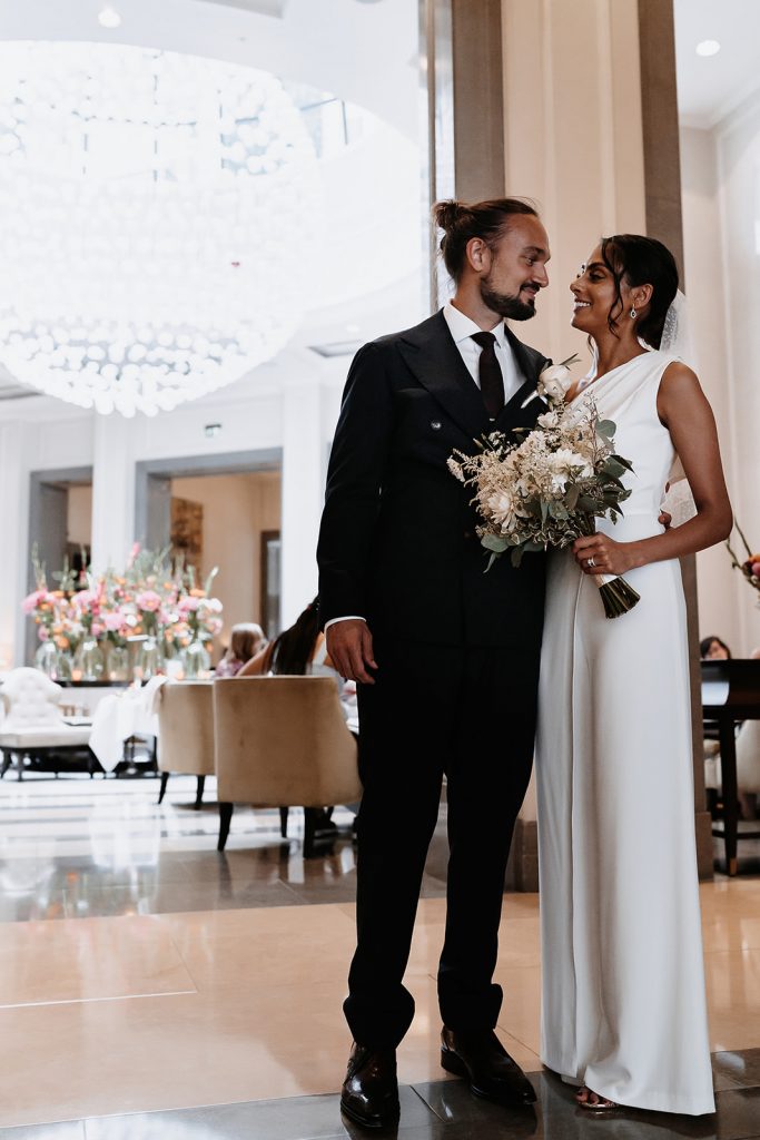 A bride and groom stand looking at one another and smiling in the grand room of the luxury hotel Corinthia London. The bride wears a chic bridal jumpsuit by Halston and holds a small wedding bouquet of greens and white flowers and foliage.