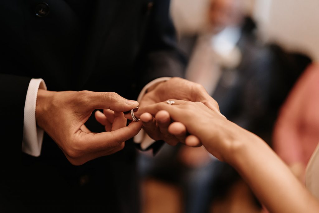 A groom gently places a wedding ring on his bride's finger. You cannot see their faces, just the torso of the man and the arm of the bride.