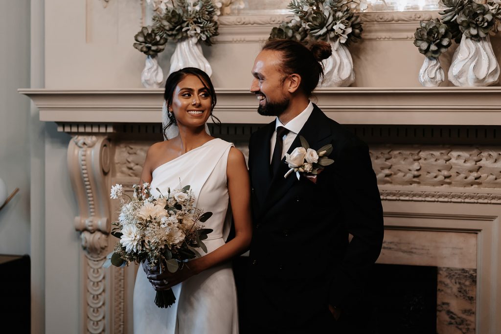 A bride and groom stand next to one another in The Old Marylebone Town Hall, London, during their intimate wedding ceremomny. The bride wears a one shouldered bridal jumpsuit and holds a beautiful green and white wedding bouquet. Her new husband wears a smart dark suit and tie with a white shirt and has his long her tied up in a top knot.