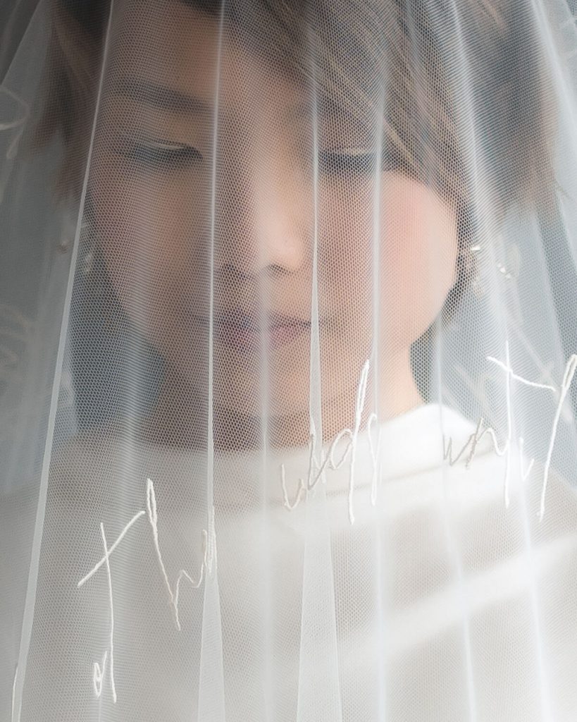A close up of a beautiful bride's face. She has short brown hair and looks down as if thinking. She has white eyeliner on her top eyelids. Over her face is a sheer, delicate veil which has script writing on it.