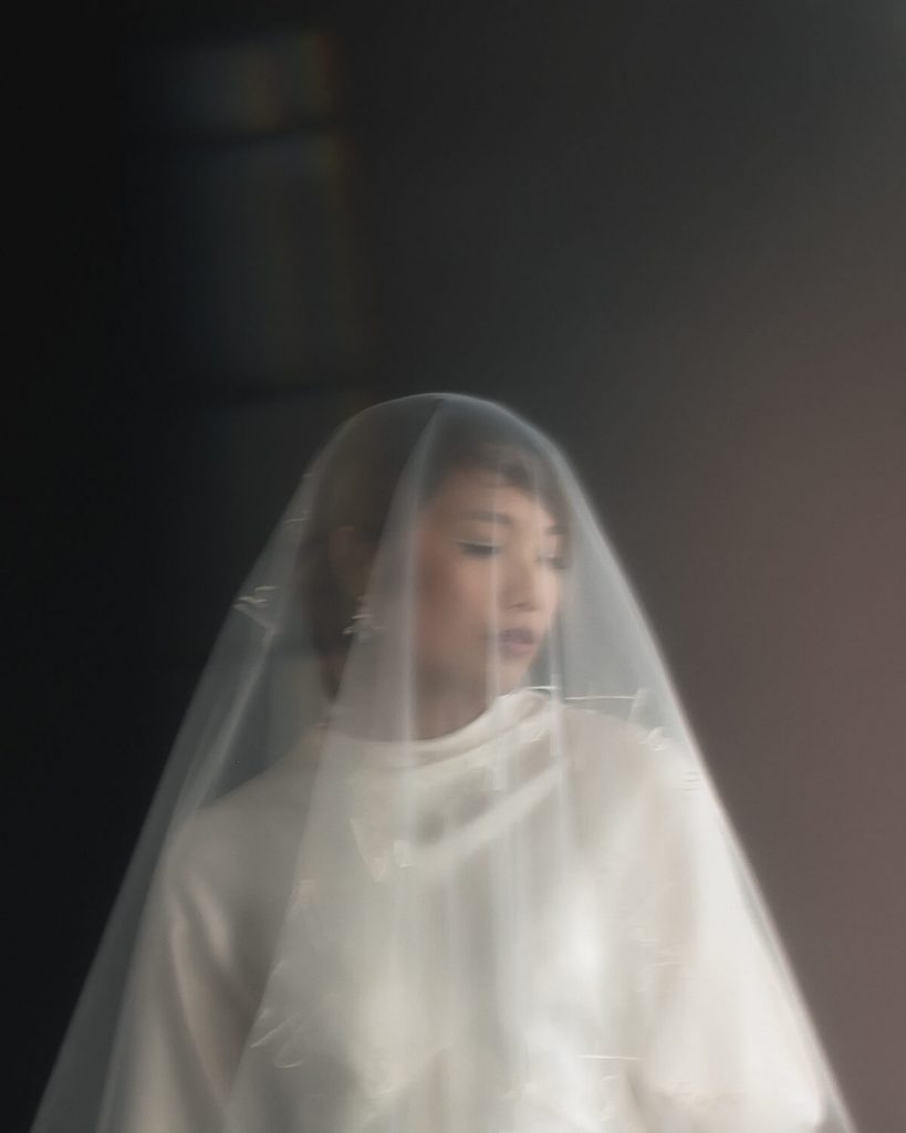 A bride with short brown hair closes her eyes and tilts her head towards the ground as if deep in thought. Covering her face is a sheer veil which has embroidered words written on it.