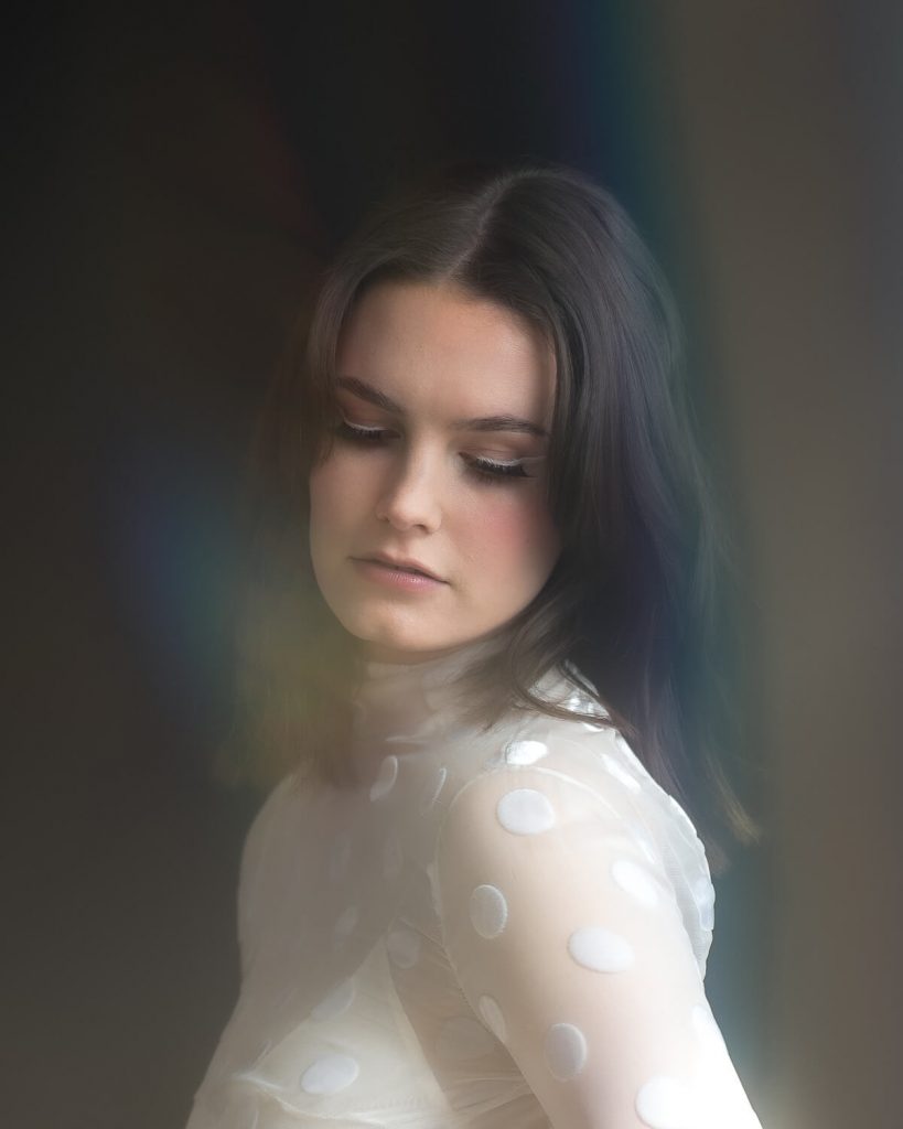 A bride wearing a high neck white polka dot wedding dress looks down towards the floor. She has shoulder length slightly wavy brown hair with a centre parting. She looks deep in thought.