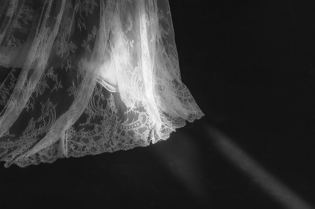 A close up of the skirt part of a wedding dress in whcih you can see a foot and part of a wedding heel. The dress is made of beautiful delicate lace and the sun is shining so warmly onto the dress.